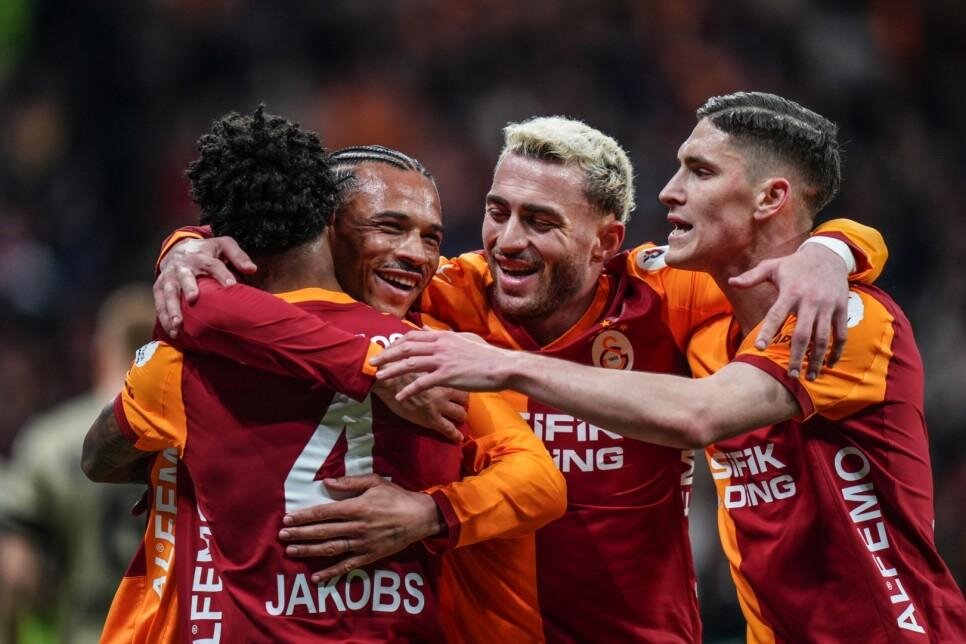 Four soccer players in orange-red jerseys embrace in celebration on the pitch; ‘JAKOBS’ is visible on one back.