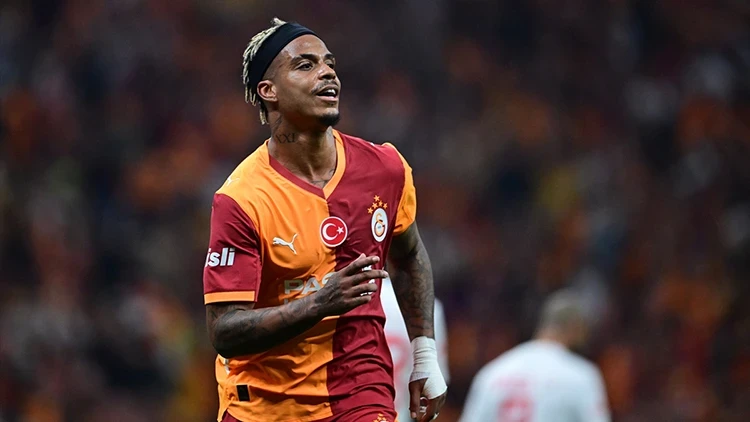 Soccer player wearing a Galatasaray jersey (orange and red) on the field, with a headband, mid-celebration.