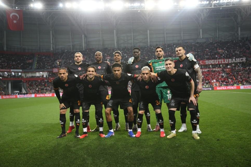 Soccer team in black uniforms posing for a group photo on a stadium field with a Turkish flag in the background.
