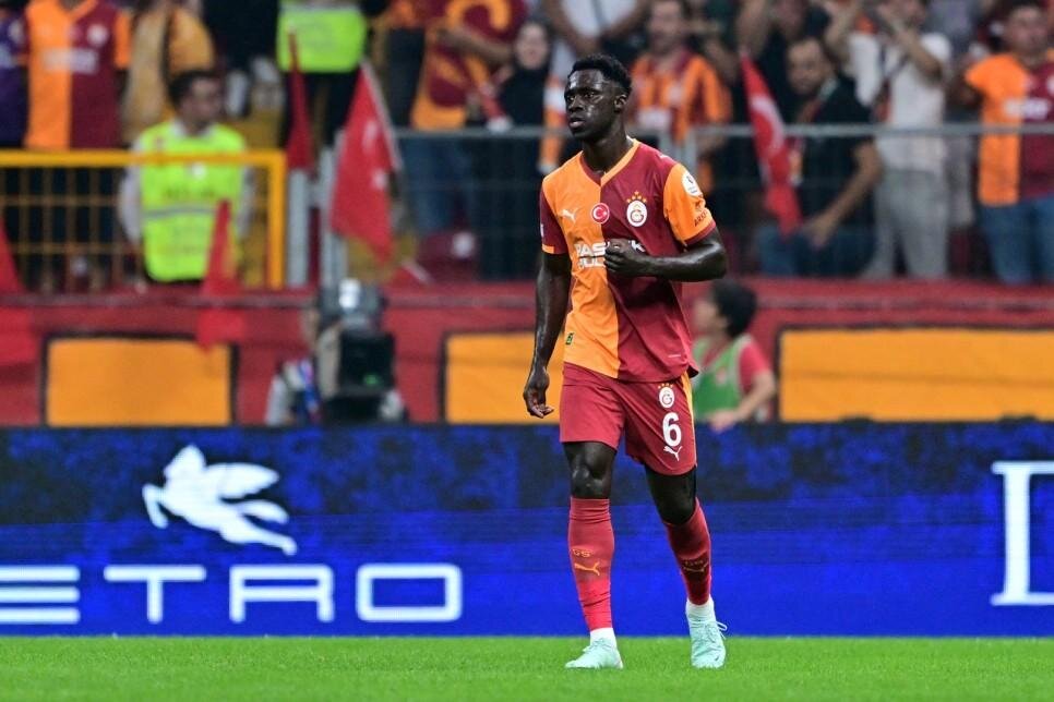 Footballer in orange-and-red Galatasaray kit walking on the pitch with cheering fans in the stands behind him, number 6 visible on shorts.