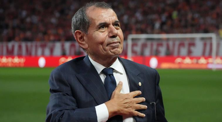 Man in a dark suit on a football field places his right hand over his heart, looking to the side as if acknowledging the crowd.