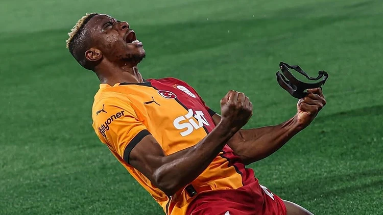 Soccer player in orange and red kit celebrating on the grass, fists clenched and mouth open in jubilation, holding a mouthguard.